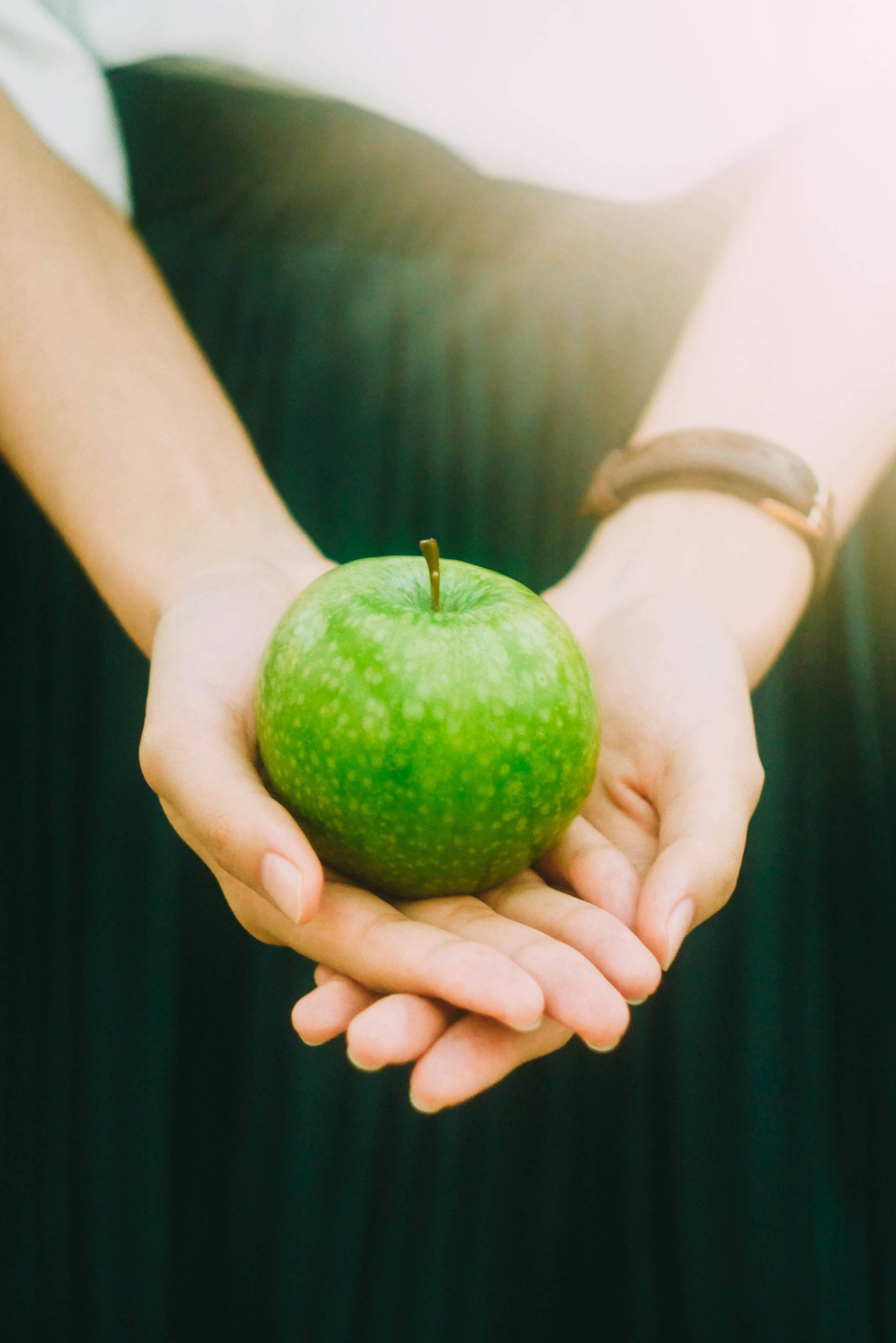 Woman wearing watch and green dress, holding green apple with sunlight streaming in