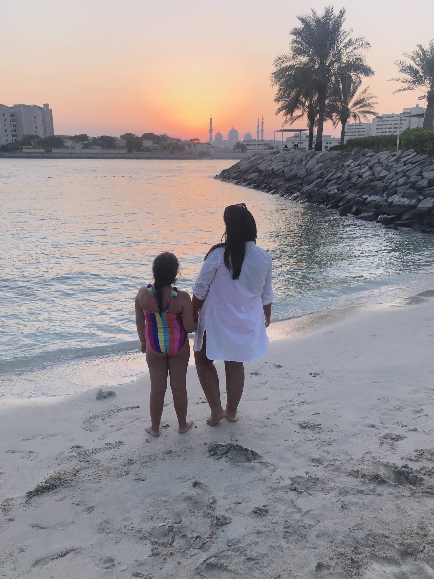 woman and child stood on beach staring at sunset over mosque
