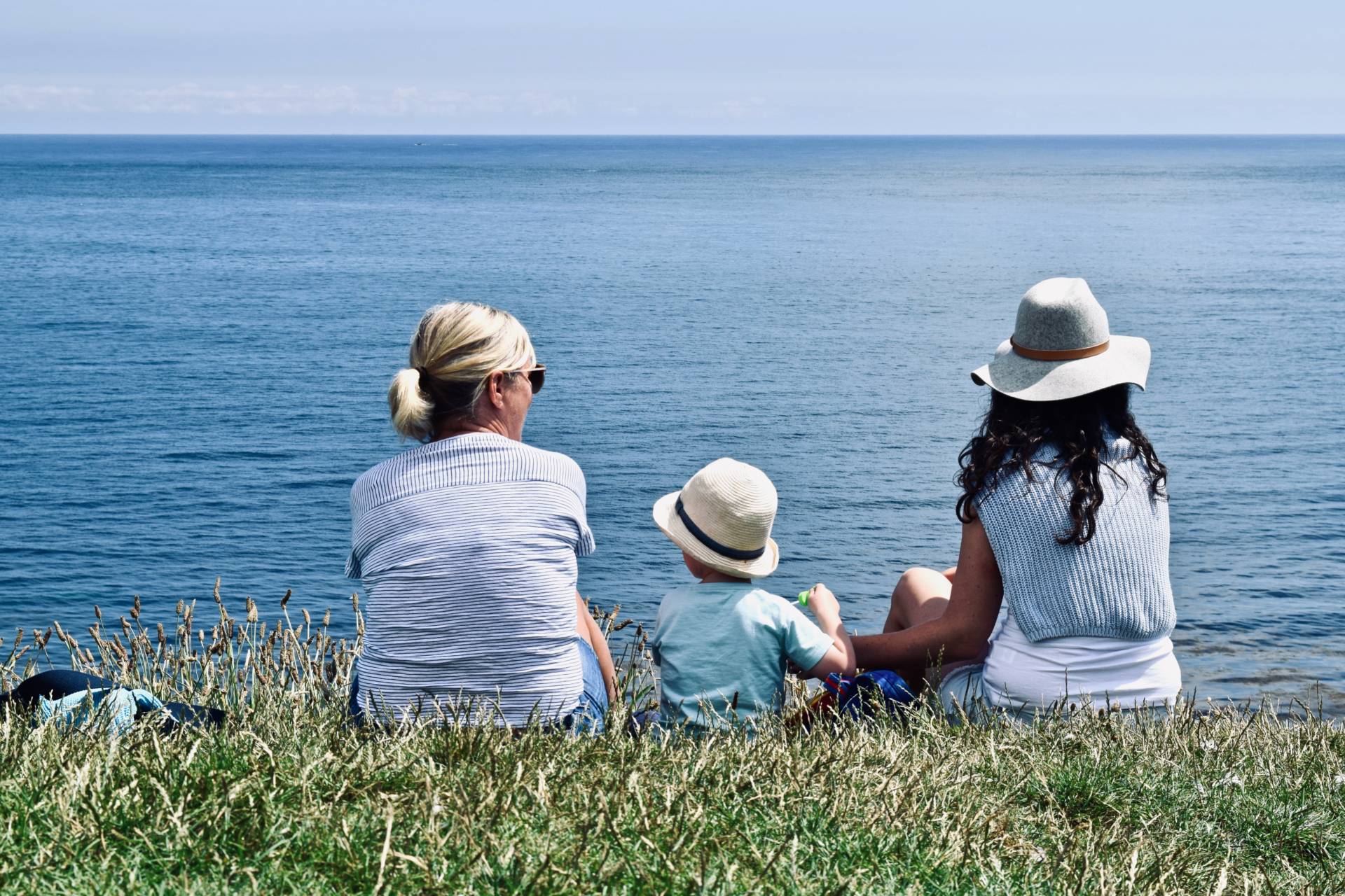 older woman, child and woman sat on grass looking out to sea.