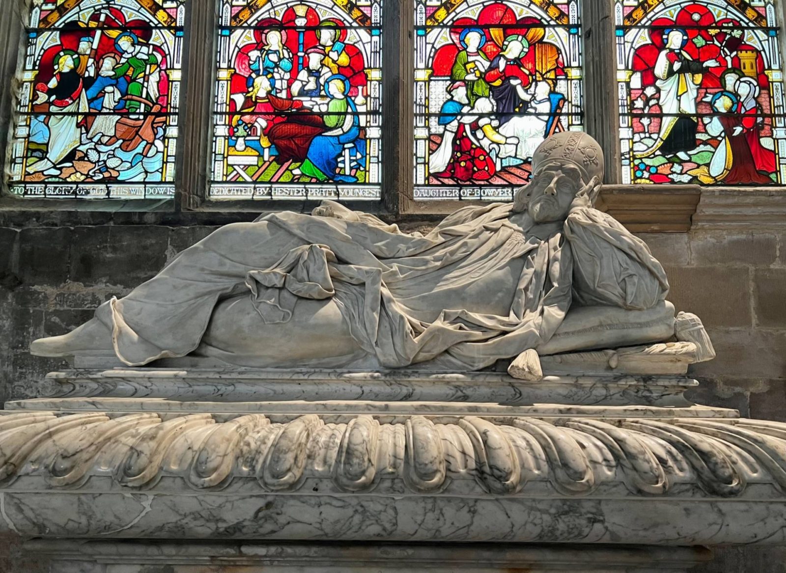 marble statue of a man laid on his side in front of stain glass windows